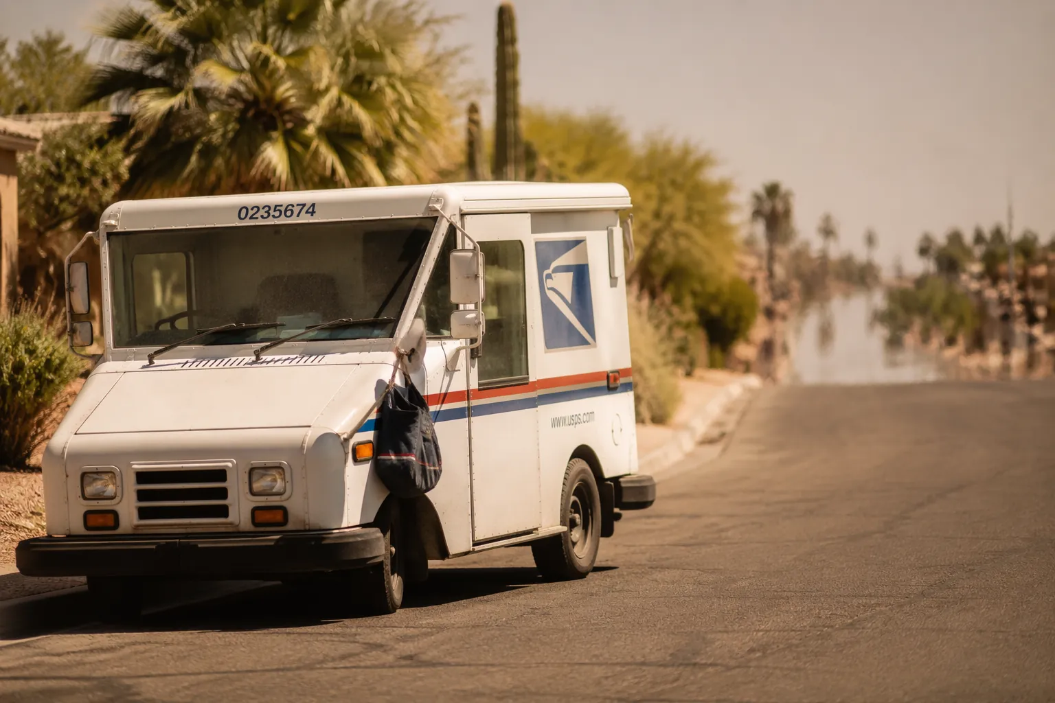 USPS-style Grumman LLV mail truck on a sun-baked residential street in oppressive heat