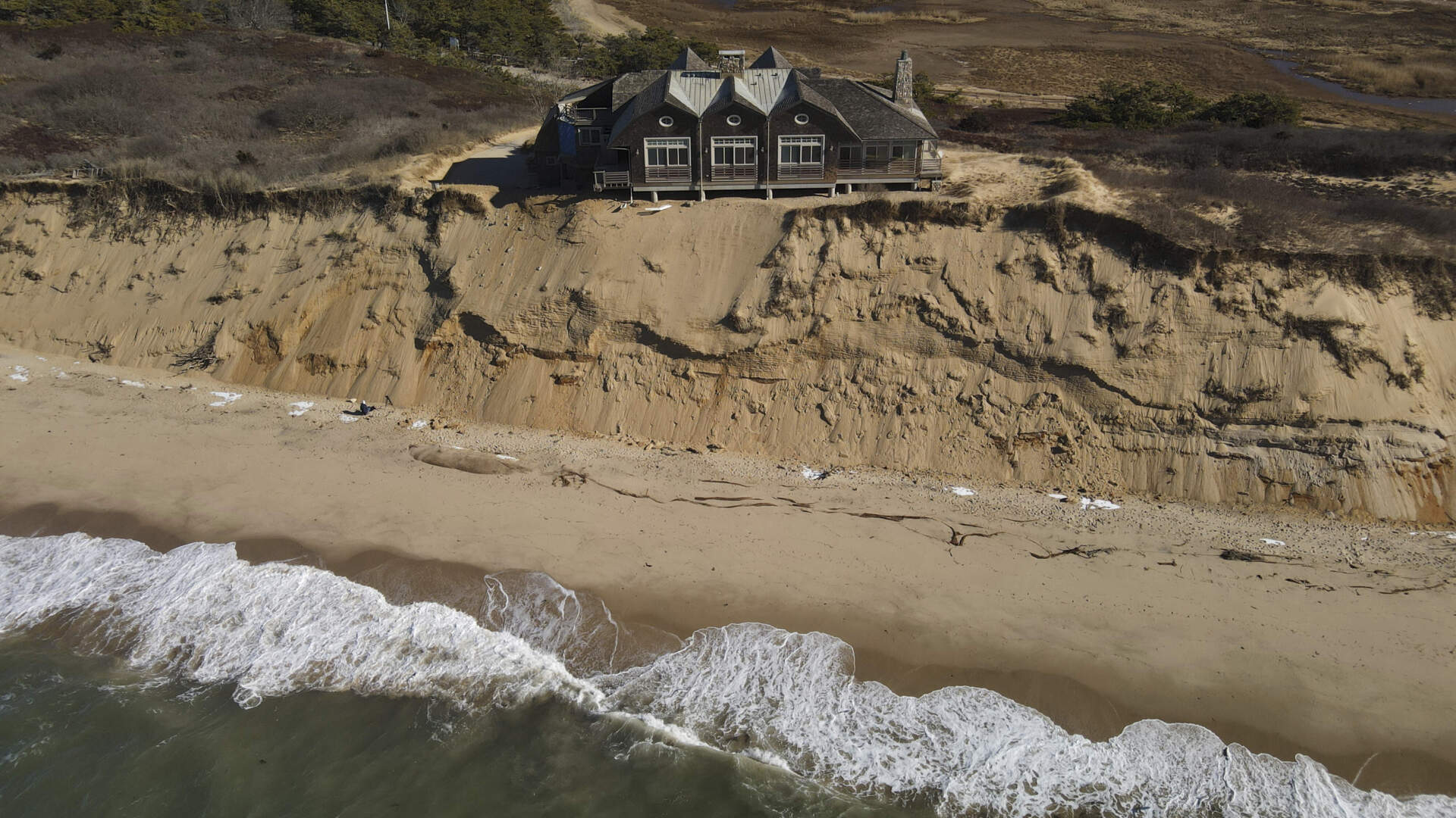 A large modern house perched at the edge of an eroding sandy coastal bluff above Cape Cod Bay