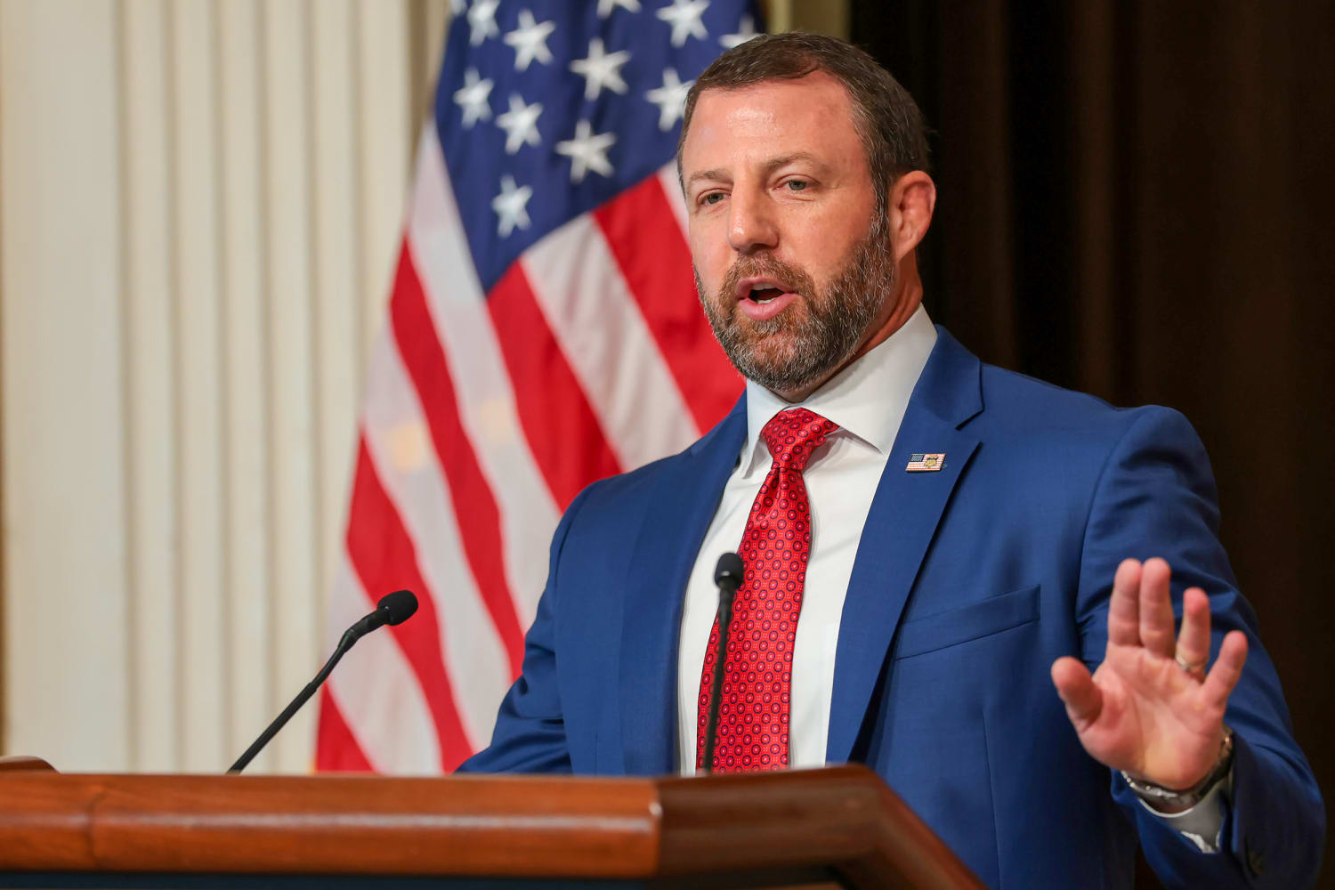 Markwayne Mullin speaking at a Justice Department press conference podium with microphones and an American flag behind him